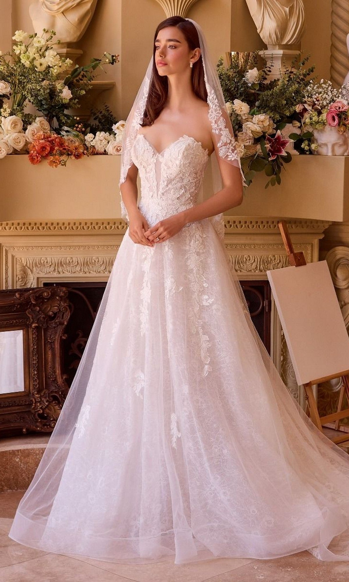 A woman stands indoors in front of a fireplace and floral arrangements wearing the Andrea and Leo WL052 Long Wedding Dress, featuring white floral lace appliqué and an elegant A-line silhouette.